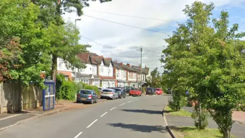 A curving residential street with trees on both sides of the road and cars along the left hand side