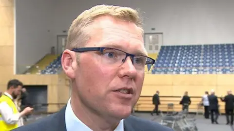 Nick Forbes has short blonde hair and wears glasses. He is standing in a sports hall after an election count.