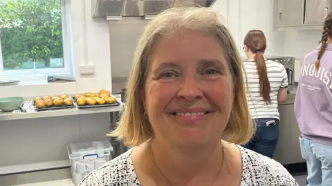 A lady with blonde/sandy hair, just above her shoulders, she has a black and white top on with a silver necklace with a small silver pendant, behind her are two ladies in a kitchen with a tray of jacket potatoes on the left on a worktop.