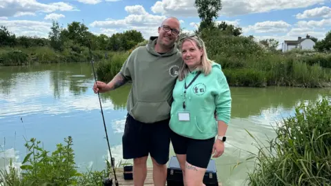 Khadijah Hasan/BBC A man and a woman wearing Recovery Rods hooded tops and black shorts stand in front of a fishing lake. The man has a fishing rod in one hand and has his other arm around the woman's back as they smile towards the camera