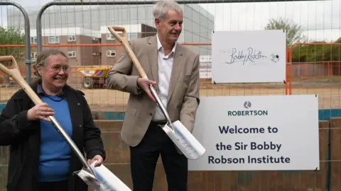 Prof Ruth Plummer and Mark Robson holding spades next to a sign pinned to the construction fencing, reading: "Welcome to the Sir Bobby Robson Institute". She has long grey hair tied back and wears glasses, a black coat and blue top. He has a beige suit jacket and white shirt and looks uncannily like his dad.