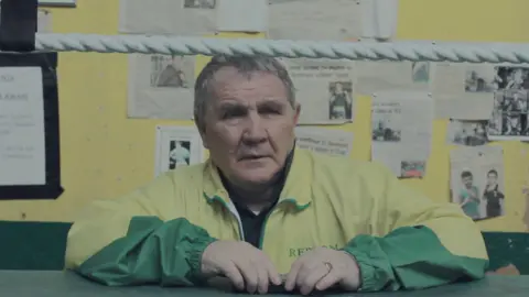 Ryan Pickard/ Learning The Ropes A man wearing a green and yellow tracksuit leans over the edge of a boxing ring. There are newspaper clippings pinned up on the yellow wall behind him.
