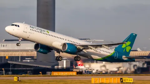 An Aer Lingus aircraft is taking off from a runway. The plane is white with patches of blue it also has a shamrock on the rear fin. 
