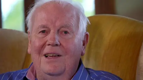 BBC A picture of Sir David Kirch. He is smiling at the camera wearing a striped blue shirt. He sits on a yellow sofa. 