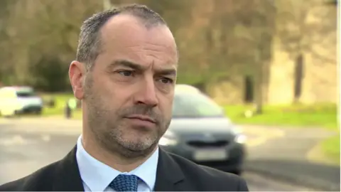 Patrick Corrigan is looking to someone beyond the camera with a neutral expression on his face. He has short black and grey hair and a stubbly beard. He is wearing a black suit jacket, a light blue collared shirt and a patterned blue tie. He is standing on a road, with some parked cars in the background but they are blurred.