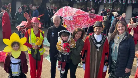 Welsh government Eluned Morgan smiles at the camera while holding a child. She is joined by the Mayor of St David's and members and children of the community. One child wears a yellow daffodil on their head. One woman is dressed red with a neon vest and has a red dragon on her back.