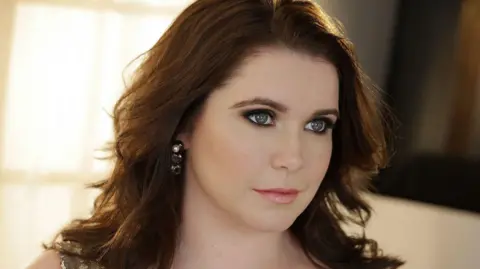 Kristin Hoebermann Tara Erraught is wearing silver earrings and her brown hair is down. She has Green eyes and is standing in front of a brightly lit background