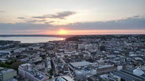 BBC A drone shot above St Helier as the sun sets into the sea beyond.