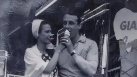 Martina Penna A young woman with long dark hair wearing a white hairband and spotty dress is holding an ice cream, that the young man next to her his licking. He is wearing a t-shirt with a collar. Next to them is a giant wooden ice cream, as tall as they are