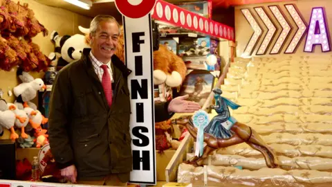 Nigel Farage, who is wearing a green wax jacket, shirt and red tie, smiles as he gestures towards a toy horse and jockey that forms part of an arcade amusement. The rider is wearing a blue Reform rosette and is headed towards a finish line. 