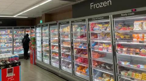 Rows of brightly coloured frozen food packets and bags stacked behind glass cabinets in a supermarket.