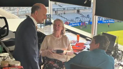 Supplied A man in a suit stood next to Jane and another person who are sat down in a cricket scoring box. Jane is holding a box with a medal in.