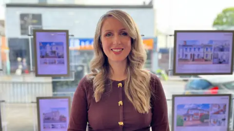 BBC News Angharad Trueman, wearing a burgundy cardigan with gold bow buttons and her long blonde hair on waves over her shoulders. She is smiling at the camera and behind her there are letting or for sale adverts in an estate agents window. 