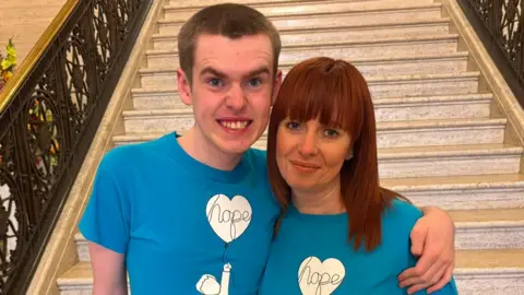 A boy with dark hair has his arm around a woman with dark hair, they are both wearing blue and white tops.