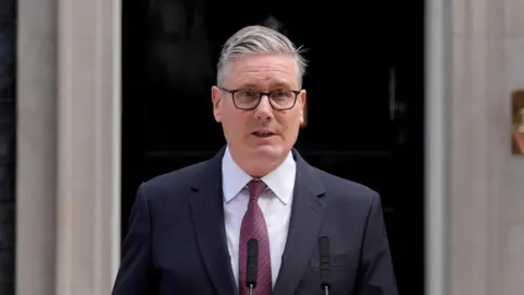 Sir Keir Starmer wearing a dark suit and tie standing outside 10 Downing Street. He is giving a speech from a lectern.