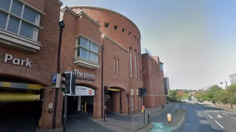 The Lanes shopping centre is a multi-storey red brick building. A sign for a car park can be seen on the left and the shop, Debenhams, can be seen on the right. In the middle, a sign reads: "The Lanes Shopping Centre".