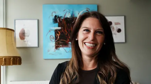 John Fairhall/BBC The head and shoulders of a woman wearing a black top, with long brown hair, while standing in front of an artwork on the wall. She is looking into the camera with a big smile.