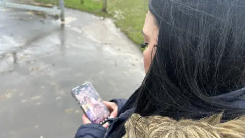 A woman looks at her phone. She has black hair. It is a wet and drizzly day. 