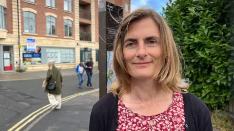 BBC/RICHARD EDWARDS A woman standing on a street in York. She is wearing a red, patterned blouse and a navy-blue cardigan. She has shoulder-length fair hair. People are crossing the road in the background behind her.