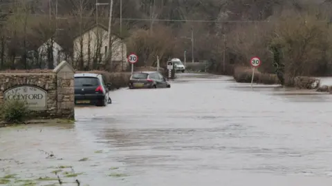 Two cars are submerged in floodwater on a road in Colyford, Devon. A sign with the village's name on it attached to a brick wall is in the foreground. A pair of 30mph signs are in the distance.