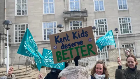LDRS A cardboard sign that reads "Kent kids deserve better" and multiple light blue flags that have the emblem of the National Education Union on them. A large, grand stone building can be seen in the background.