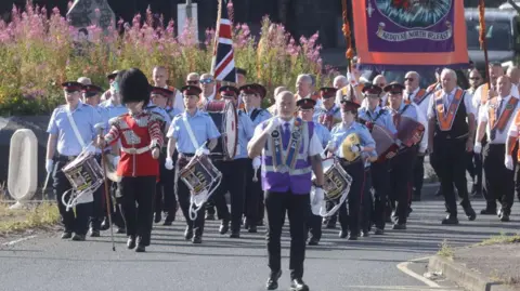 Pacemaker Orange Order and band members marching with a banner saying on top Lol Earl of Erne 647 which features a person beside a cross and below it the words Ardoyne North Belfast. The people marching with the band are carrying drums and accordions.