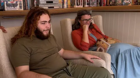Solly and Amy are sitting in lounge chairs looking at a tv out of frame.