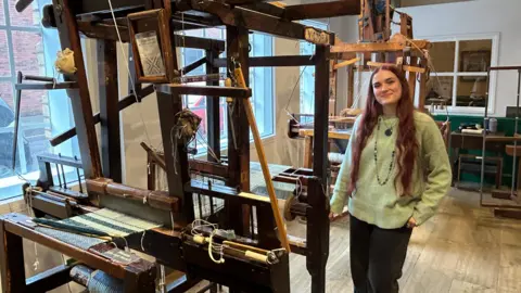 Charlotte Blazier stands next to one of the two Victorian handlooms. The machine is made of wood and there is half-finished fabric on it. 