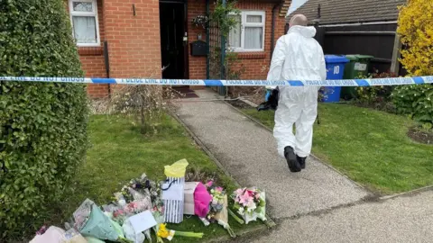PA Media The garden of a brick-built house with bushes to the side and small grass lawns either side of a stone path. There is police tape across the garden, and a person in a white hazmat overall is walking towards the front door. There are bouquets of flowers on the lawn to the left.