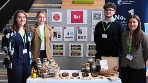 Harper Adams University The three women and one man stand by a table with their snacks and product information on. A board is visible behind them with small posters one and they are stood by some stairs to the left.