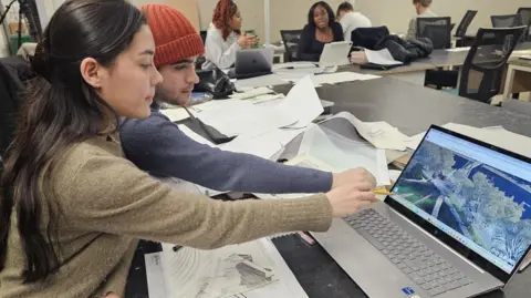 Students are sitting around a desk with sketches and paper scattered over the table. A woman with long brown hair and a man with an orange woolly hat are in the forefront of the picture pointing to a computer screen. 