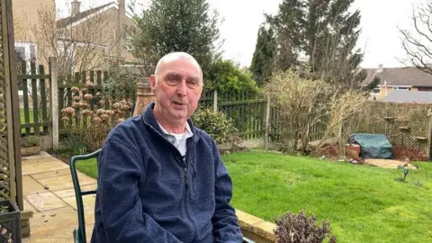 Jonny Humphries/BBC Steve Cervi, 71, who has a bald head with grey hair on the sides, is clean shaven and is wearing a blue fleece over a grey polo-neck t-shirt, sitting on a metal garden chair in front of a patio and a grassy lawn. 