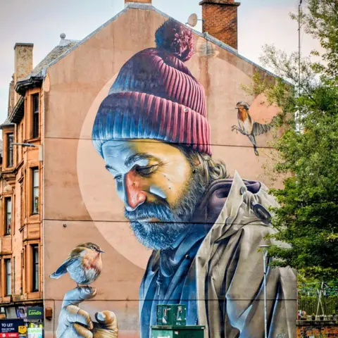 Nicolas Raes The St Mungo mural on a gable end in Glasgow. It features a man with a dark beard wearing a red bobble hat allowing a robin to perch on the end of his finger. A green tree is partially covering the right side of the image.