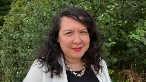 Martin Heath/BBC Head-and-shoulders portrait of Victoria Collins standing outdoors in front of dense green foliage. Collins has dark, shoulder-length curly hair and is wearing a light-coloured blazer over a black top with a chunky chain necklace. The image is taken in daylight with natural lighting, and the leafy background fills the frame behind the subject.