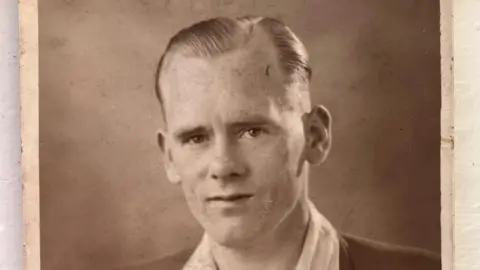 Andrea Morrison A sepia-toned portrait photograph shows an individual dressed in a formal suit jacket with wide lapels over a light-colored collared shirt. The jacket has a small pin on the lapel and a pocket on the left side. The photo has worn edges, indicating age, and is placed on a light surface.