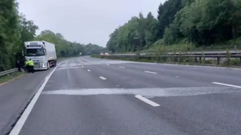 An empty three-lane motorway with one white lorry parked on the hard shoulder. There are damp patches across the motorway and trees lining the edges