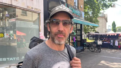A man with a grey shirt, hat and beard stands on a high street.