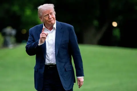 Trump raising an index finger and smiles as he walks on a green lawn. He is wearing a navy suit.