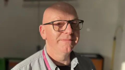 Chris Priest wearing a grey shirt and large dark brown glasses. He has a pink lanyard around his neck