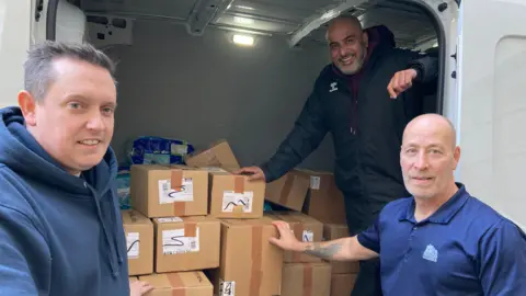 Three men stand at the back of a van which is loaded with several brown cardboard boxes and large plastic packets.