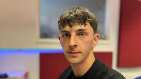 Alex Bish/BBC A young man with brown hair looks straight at the camera. He is in an office with red panels on the wall