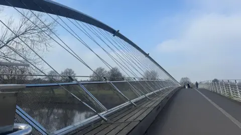 BBC/ Emily Johnson A modern pedestrian bridge with a curved, cable‑supported design rises gently over trees and water, with metal railings and mesh panels lining the walkway as a small group of people walks in the distance beneath a bright, lightly clouded sky.