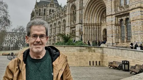 Donald Mitchell, who has grey hair and glasses stood outside a grand building wearing a green jumper and brown jacket.