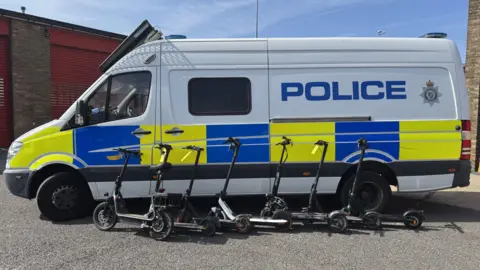 Lincolnshire Police Seven e-scooters lined up in a row against a police van. The van is white with blue and yellow markings along the middle and above it is 'POLICE' written in blue type and the police crest logo. It is parked in front of a single-storey building with red shutters down.