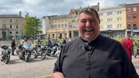 Cashmore wearing a black shirt and collar. He is smiling and standing in front of a row of bikes. 