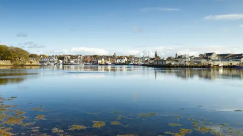 A view of Stornoway in Lewis over a calm area of sea. There are rows of buildings along the opposite shore. It is a bright sunny day and there is hardly a cloud in the blue sky.
