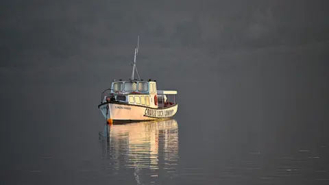 A small white boat sits still on calm, reflective water in soft evening light, with faint hills forming a dark backdrop.