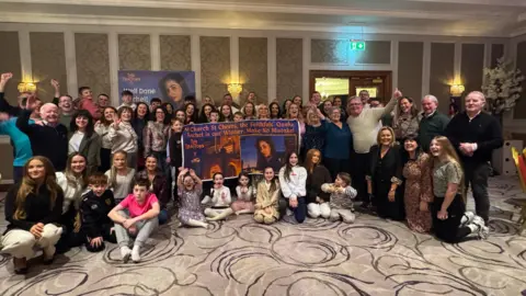 Multiple people pose for a photo in a cream coloured room. Some are waving their hands and others holding signs saying "Rachel is our winner"