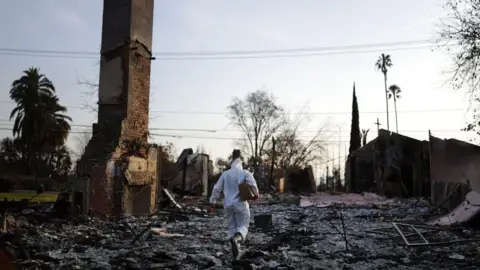 Getty Pictures A person in a protective suit walks the consequences of La -i fires. Chimney racks surrounded by debris and charred remains of buildings 