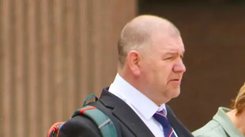 Lynda Roughley A side on view of Ian Ashworth, who has a shaven head and light stubble, walking away from court wearing a dark suit with a white shirt and blue and red striped tie.